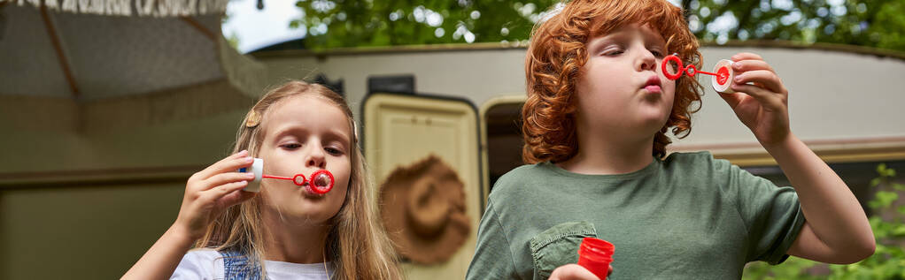 entzückende Kinder pusten Seifenblasen in der Nähe des Wohnwagens im Freien, Geschwister spielen zusammen, Banner - Foto, Bild