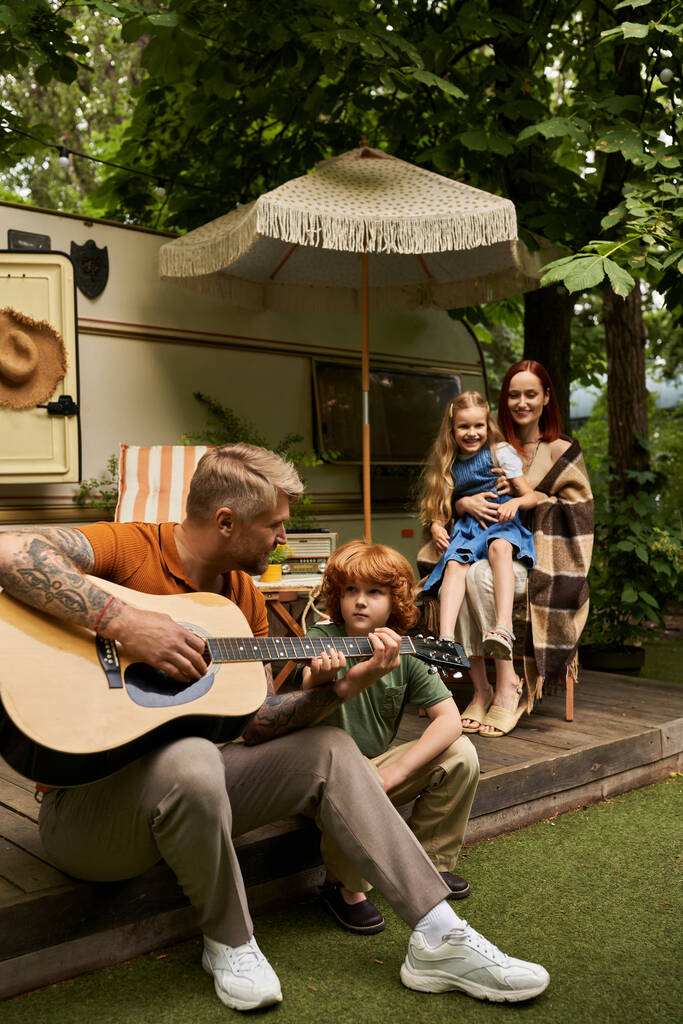 tattooed man playing guitar to attentive redhead son and family smiling next to trailer home - Photo, Image