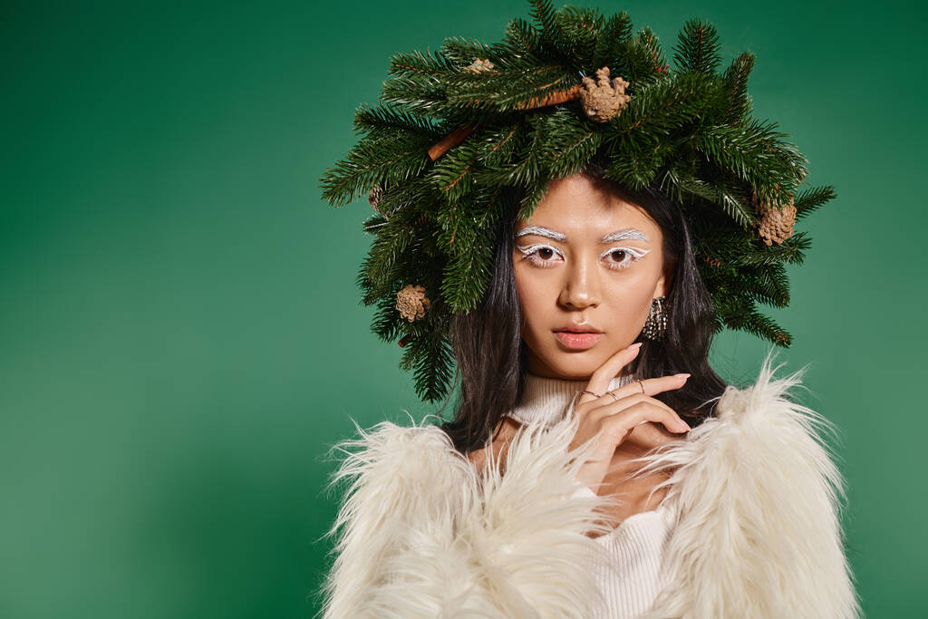 attractive woman in faux fur jacket and natural wreath on head looking at camera on green backdrop - Photo, Image