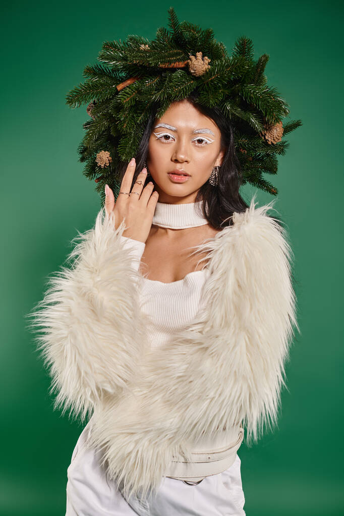 Kışın sahte kürk ceketli Asyalı kadın ve başında doğal çelenk yeşil arka plandaki kameraya bakıyor. - Fotoğraf, Görsel