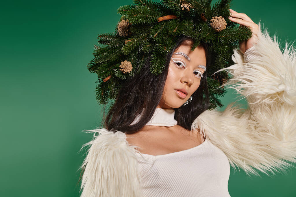 slavnostní sezóna, hezká žena s bílým make-upem a zimní oblečení pózující v věnci na zeleném pozadí - Fotografie, Obrázek