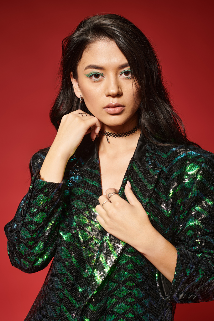 красивая азиатка в стильной зеленой куртке с блестками, смотрящая на камеру на красном фоне - Фото, изображение