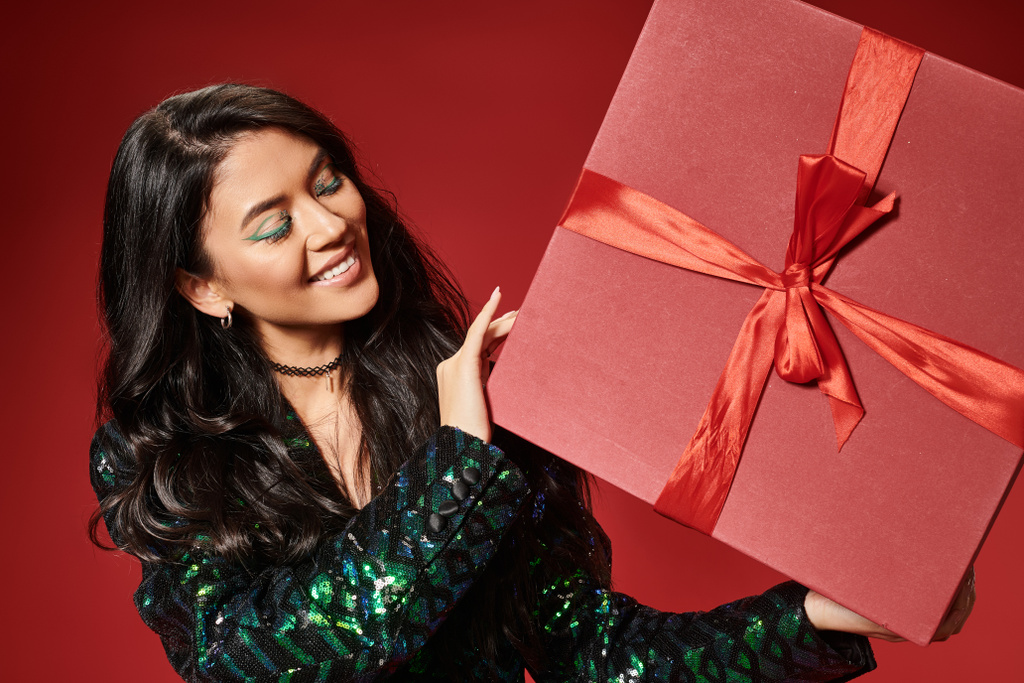smiling asian woman in jacket with sequins and blue pantyhose holding present on red backdrop - Photo, Image