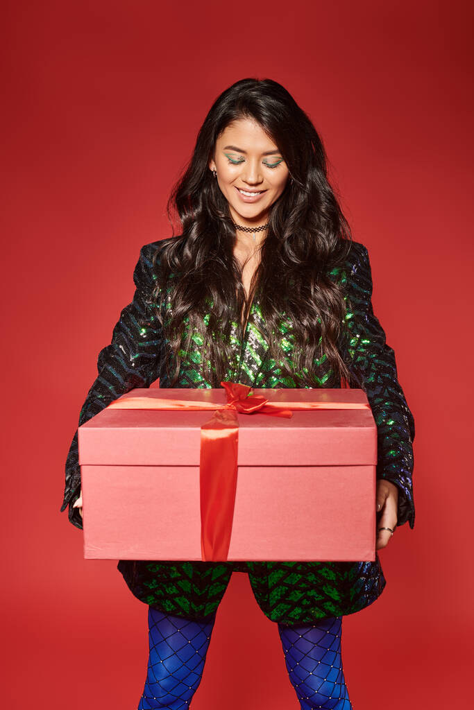 joyeuse femme asiatique en veste verte avec des paillettes regardant boîte cadeau sur fond rouge, Joyeux Noël - Photo, image