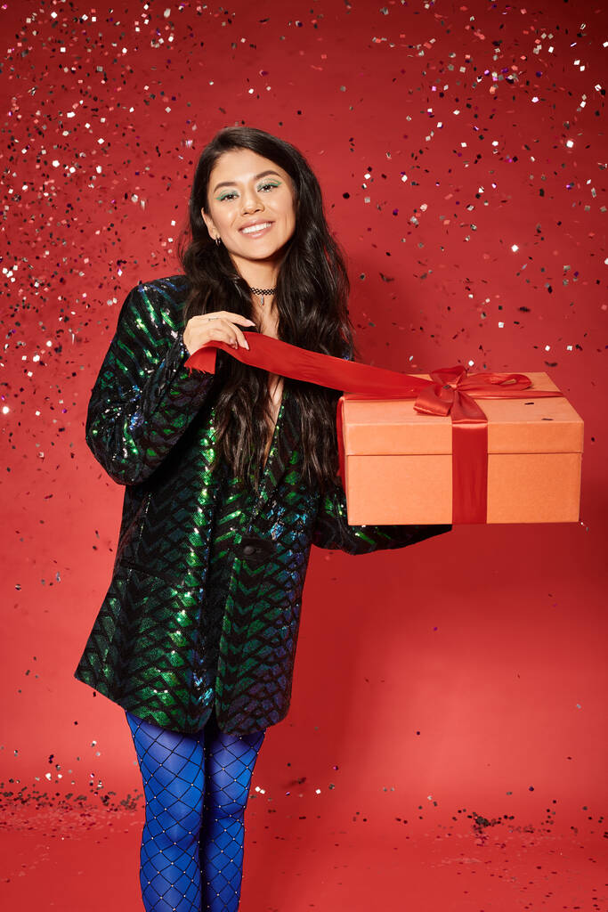 Merry Christmas, cheerful asian woman in green jacket with sequins holding wrapped present on red - Photo, Image