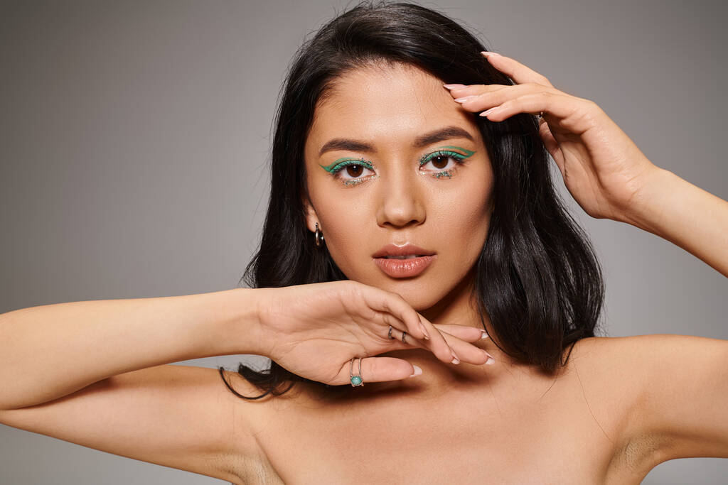 hermosa mujer asiática con brillante maquillaje ojo verde y hombros desnudos posando sobre fondo gris - Foto, imagen