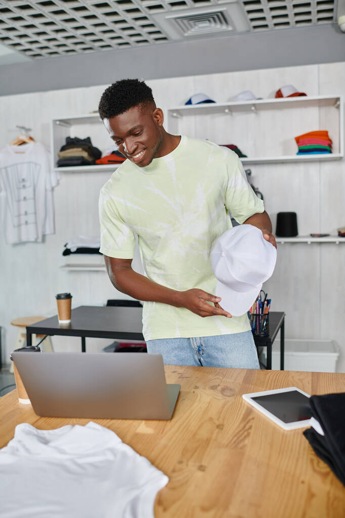 szczęśliwy afrykański projektant pokazuje biały kapelusz podczas rozmowy wideo na laptopie w studio druku - Zdjęcie, obraz