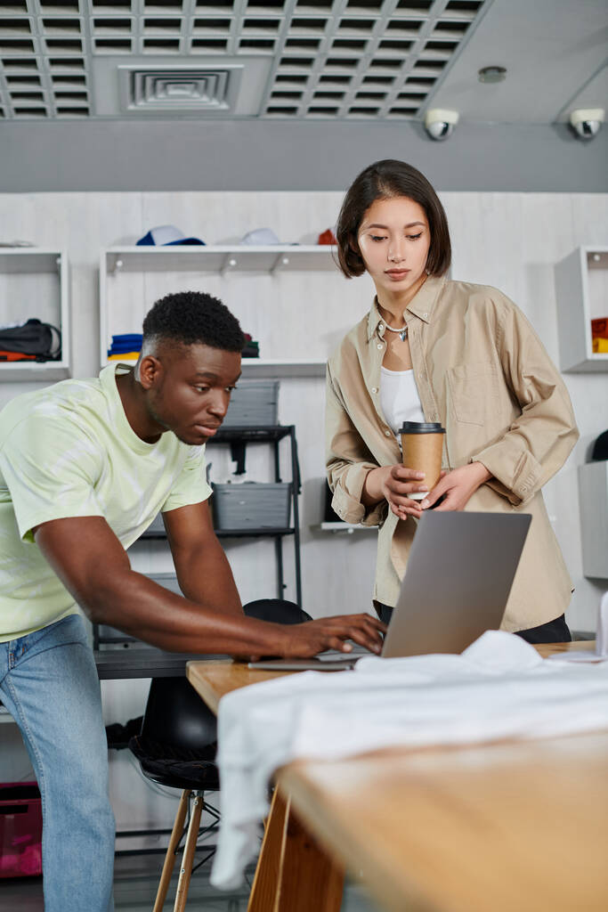 inspired african american man using laptop near asian woman with paper cup in clothing design studio - Photo, Image