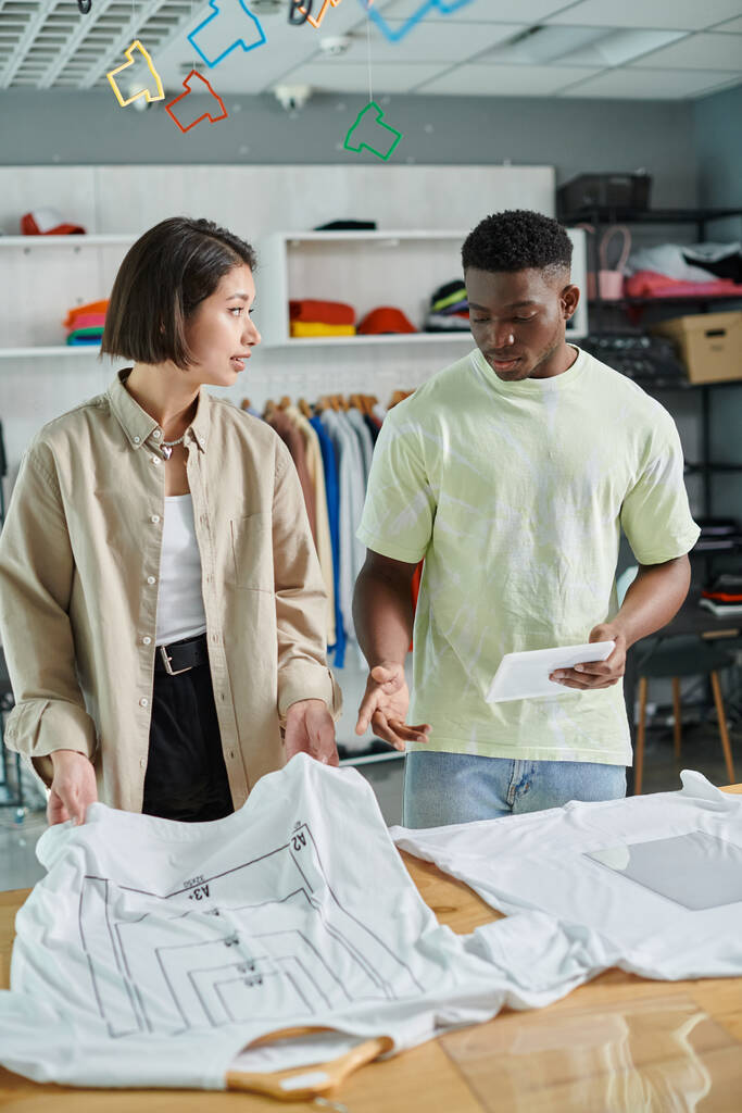 jonge interraciale kleine ondernemers praten in de buurt van t-shirts met formaat maten in print studio - Foto, afbeelding