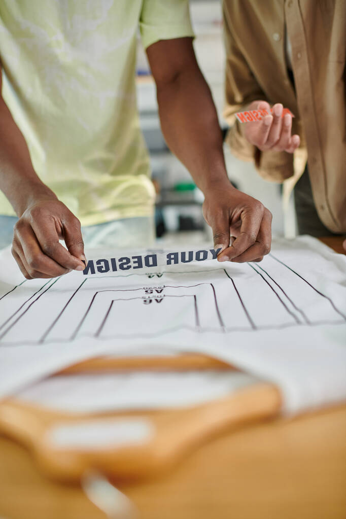 vista recortada de diseñadores de moda interracial que sostienen plantillas de texto cerca de la camiseta con tamaños de formato - Foto, imagen