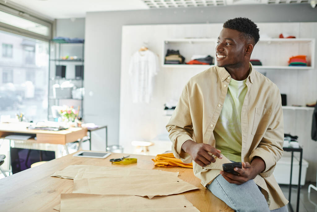 iloinen afrikkalainen amerikkalainen yrittäjä istuu älypuhelimen kanssa ja katsoo pois lähellä ompelukuvioita - Valokuva, kuva