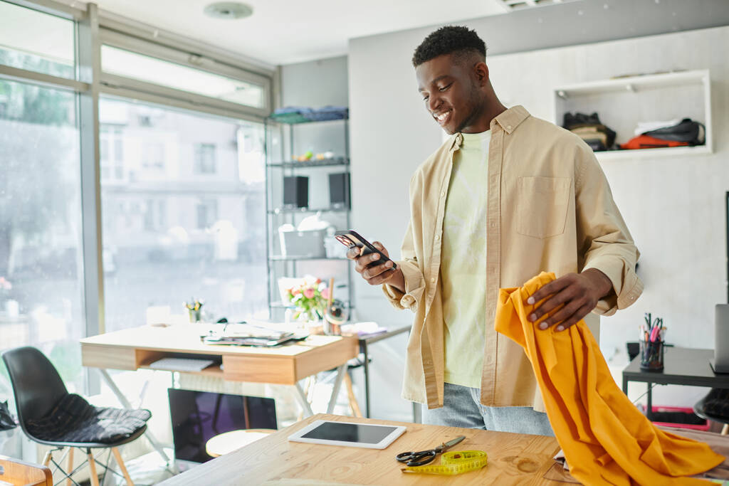 lächelnd blickt ein afrikanisch-amerikanischer Designer auf sein Smartphone, während er im Druckstudio Stoffmuster hält - Foto, Bild