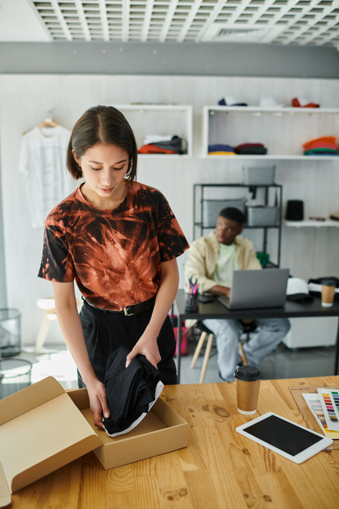 asiatische Frau beim Einpacken von Kleidungsstücken in Karton in der Nähe eines afrikanisch-amerikanischen Designers am Laptop im Druckatelier - Foto, Bild