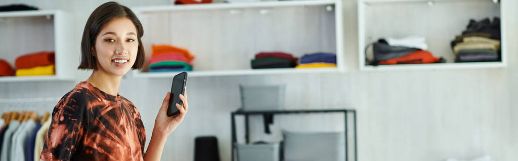 joyeux asiatique couturier avec téléphone mobile regardant caméra en textile imprimer studio, bannière - Photo, image