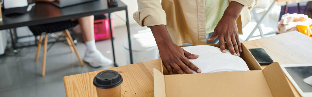 vista recortada del hombre afroamericano empacando ropa en caja de cartón cerca de taza de papel en atelier, pancarta - Foto, imagen