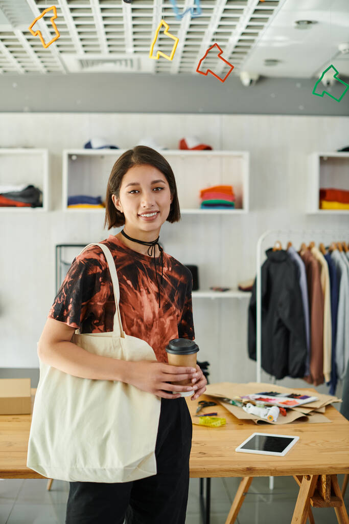 szczęśliwy azjatycki projektant z płótna torba na zakupy i kawa iść patrząc na aparat fotograficzny w studio druku - Zdjęcie, obraz