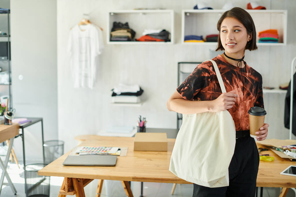 задоволений азіатський дизайнер з полотном сумка для покупок і паперовий стаканчик, дивлячись далеко в друкованій студії - Фото, зображення