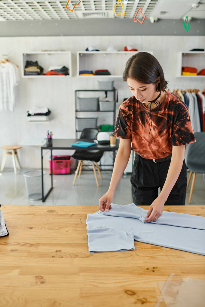 молодая и стильная азиатская женщина складывает одежду на столе в типографии, индустрии моды - Фото, изображение