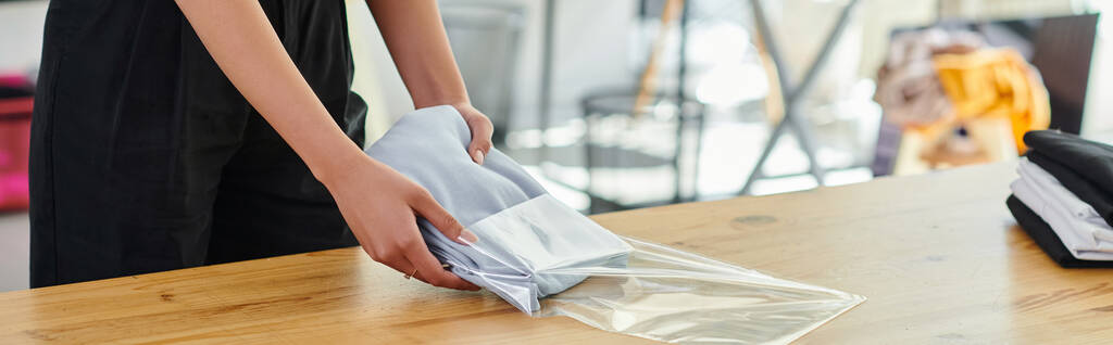 bijgesneden weergave van vrouwelijke werknemer verpakking gevouwen kleding in plastic zak op tafel in print studio, banner - Foto, afbeelding