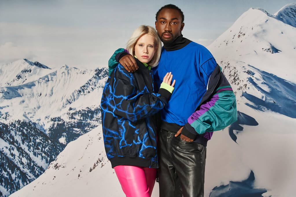 casal interracial atraente em roupas elegantes abraçando e olhando para a câmera, conceito de inverno - Foto, Imagem