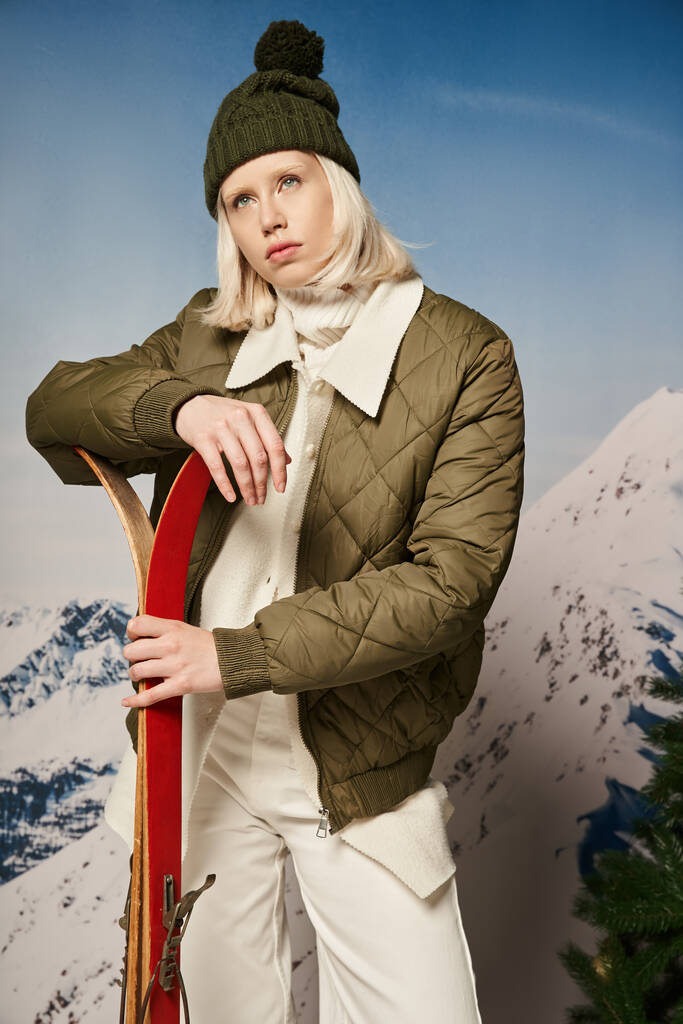 elegante donna bionda in giacca con bobble hat e sci in mano distogliendo lo sguardo, concetto invernale - Foto, immagini