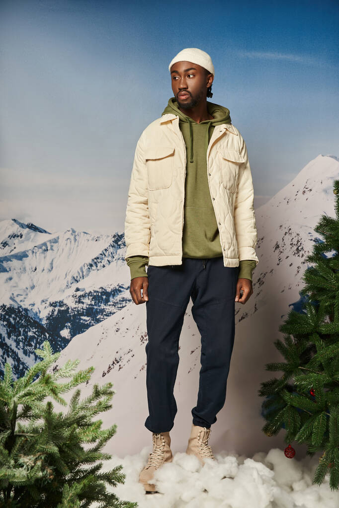 Ceketli ve bereli yakışıklı Afro-Amerikan adam köknar ağaçlarının yanında duruyor, kış konsepti. - Fotoğraf, Görsel