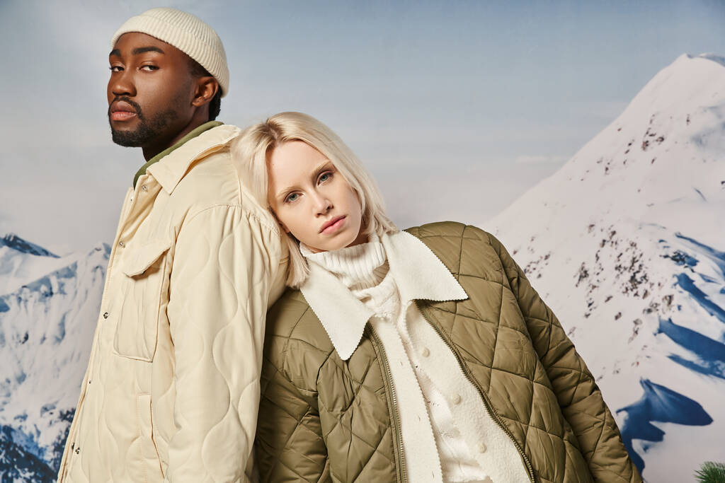 pareja interracial joven en ropa de abrigo con fondo de montaña mirando a la cámara, concepto de invierno - Foto, imagen