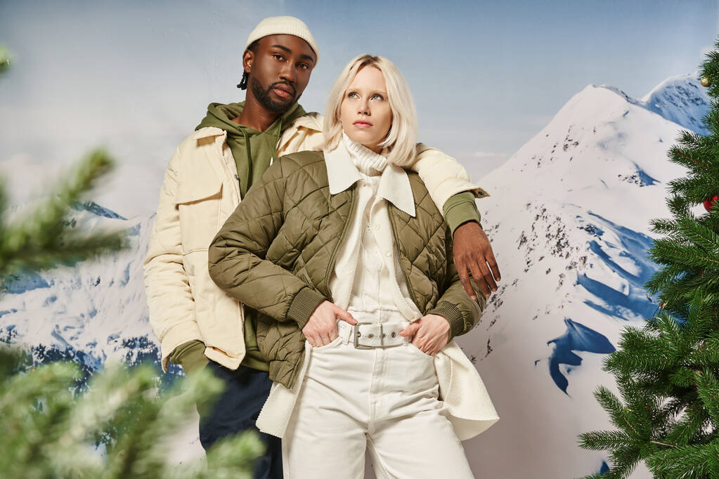 elegante pareja diversa con la montaña en el telón de fondo, brazo en el hombro, las manos en los bolsillos, concepto de invierno - Foto, imagen