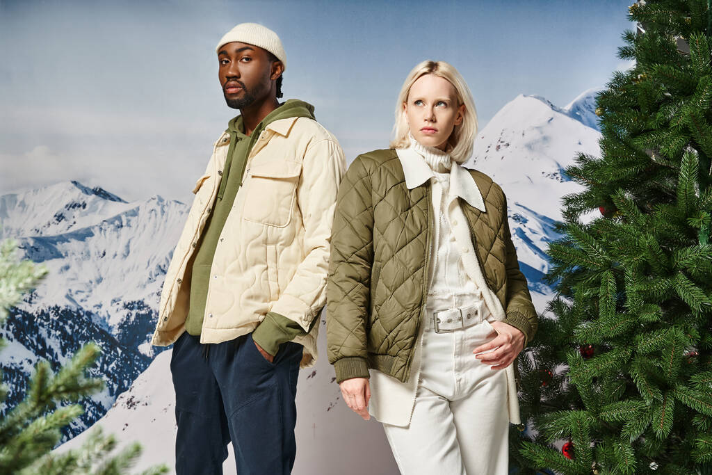 beautiful stylish couple wearing warm modish jackets and posing next to fir trees, winter concept - Photo, Image