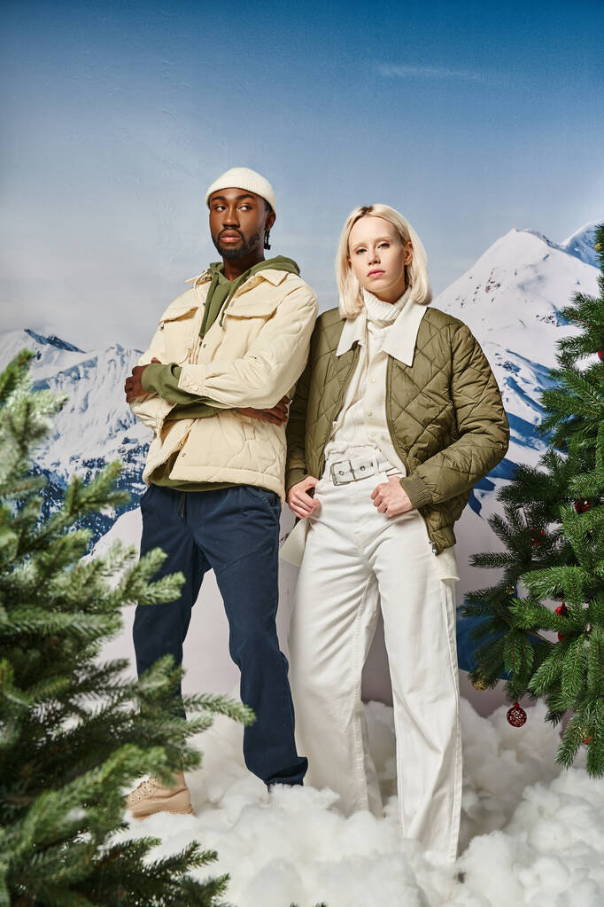 pareja de moda en traje cálido posando junto a abetos con telón de fondo de montaña, pareja diversa - Foto, imagen