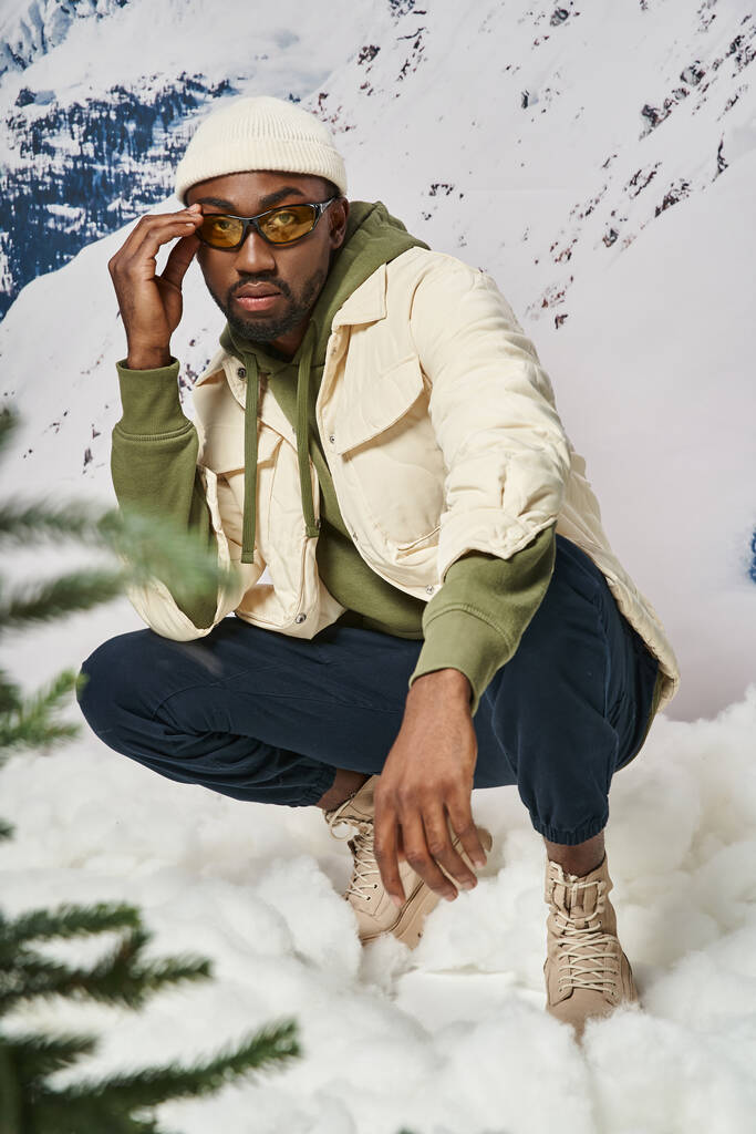 schöner stylischer Mann in warmer modischer Kleidung, der in der Hocke sitzt und seine Sonnenbrille anfasst, winterliches Konzept - Foto, Bild