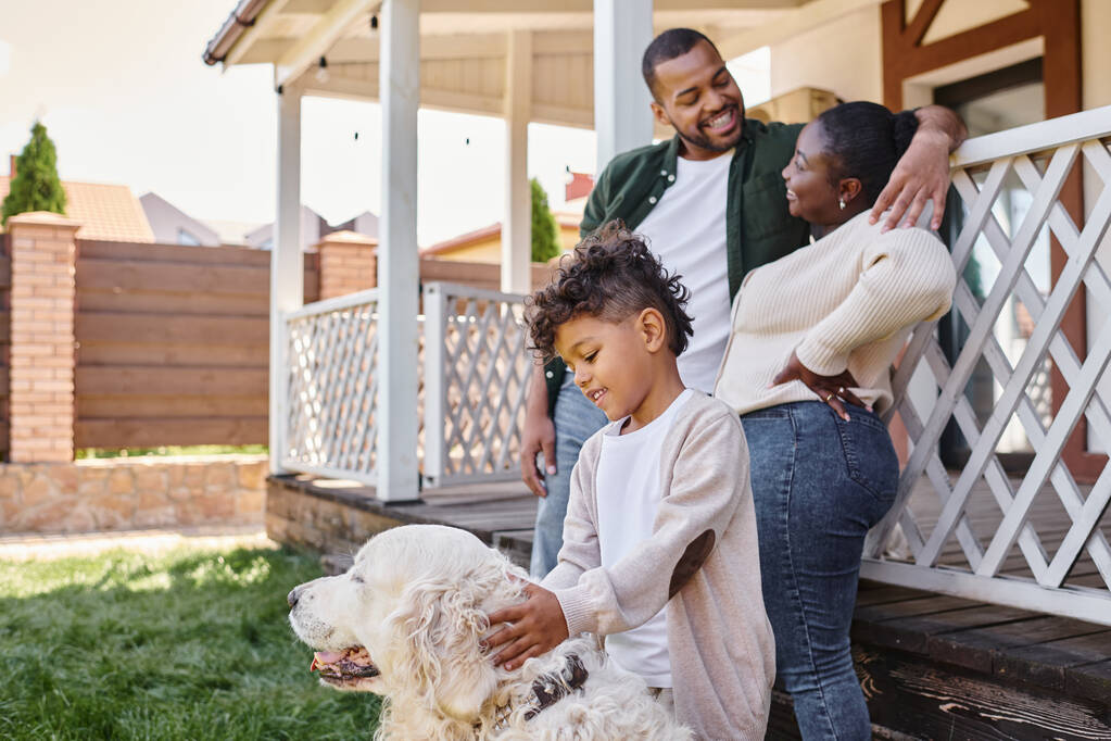 cheerful african american kid playing with dog near parents on backyard of their house, quality time - Photo, Image