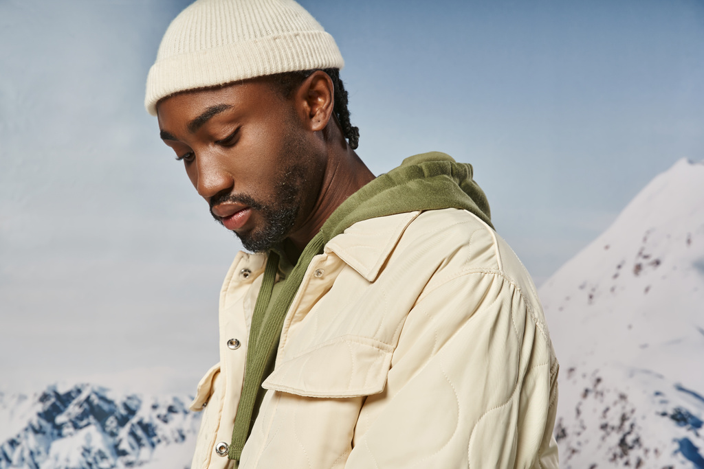 bel homme afro-américain en veste et bonnet regardant vers le bas avec fond neigeux, mode d'hiver - Photo, image
