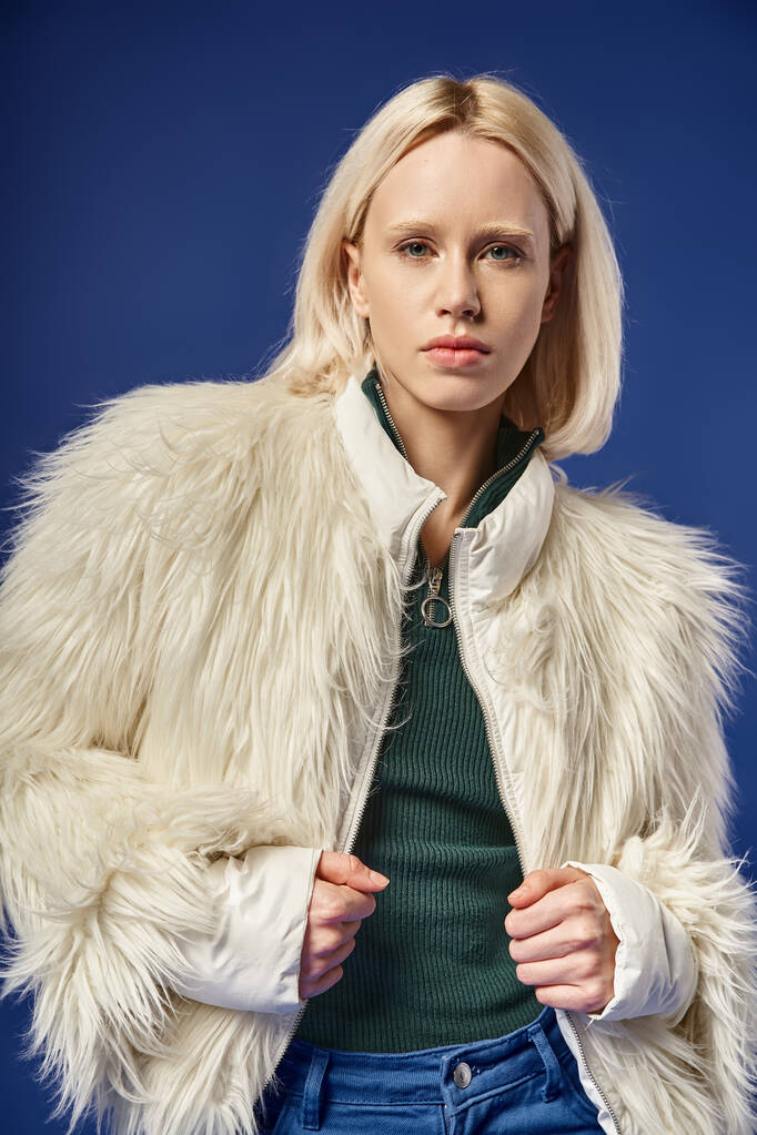 winter fashion, stylish blonde woman in faux fur jacket and denim jeans posing on blue backdrop - Photo, Image