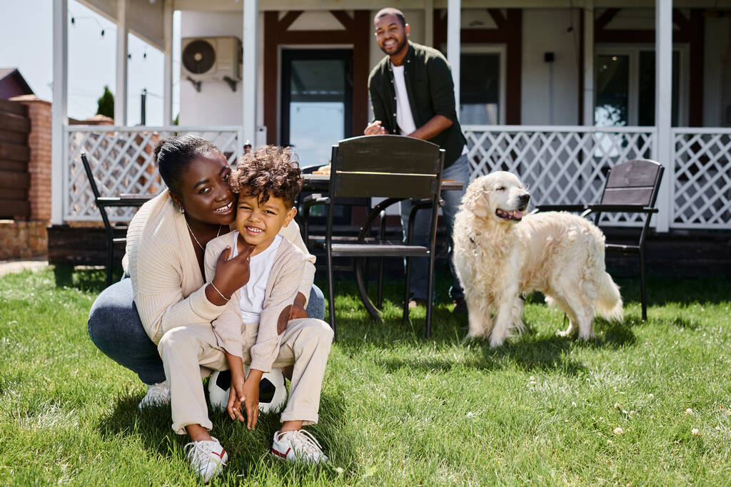 Χαρούμενη Αφροαμερικανή μητέρα αγκαλιάζει το γιο κάθεται στο ποδόσφαιρο κοντά στο σύζυγο στην πίσω αυλή του σπιτιού - Φωτογραφία, εικόνα