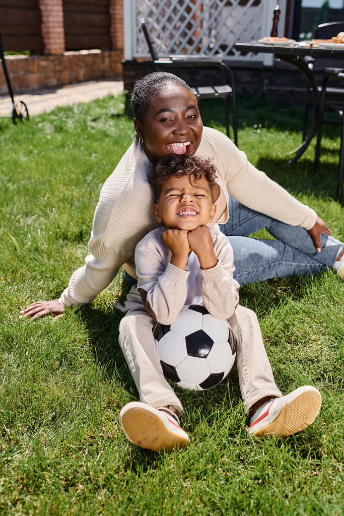 joyeuse mère afro-américaine assise sur la pelouse avec son fils à côté du ballon de football sur la cour arrière de la maison - Photo, image