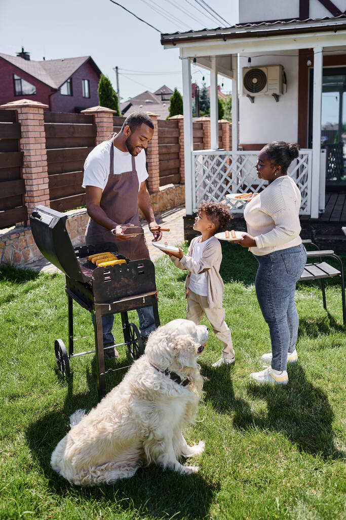 Χαρούμενος Αφροαμερικάνος που μαγειρεύει καλαμπόκι στα κάρβουνα κοντά σε σκύλο, γυναίκα και γιο στην πίσω αυλή. - Φωτογραφία, εικόνα