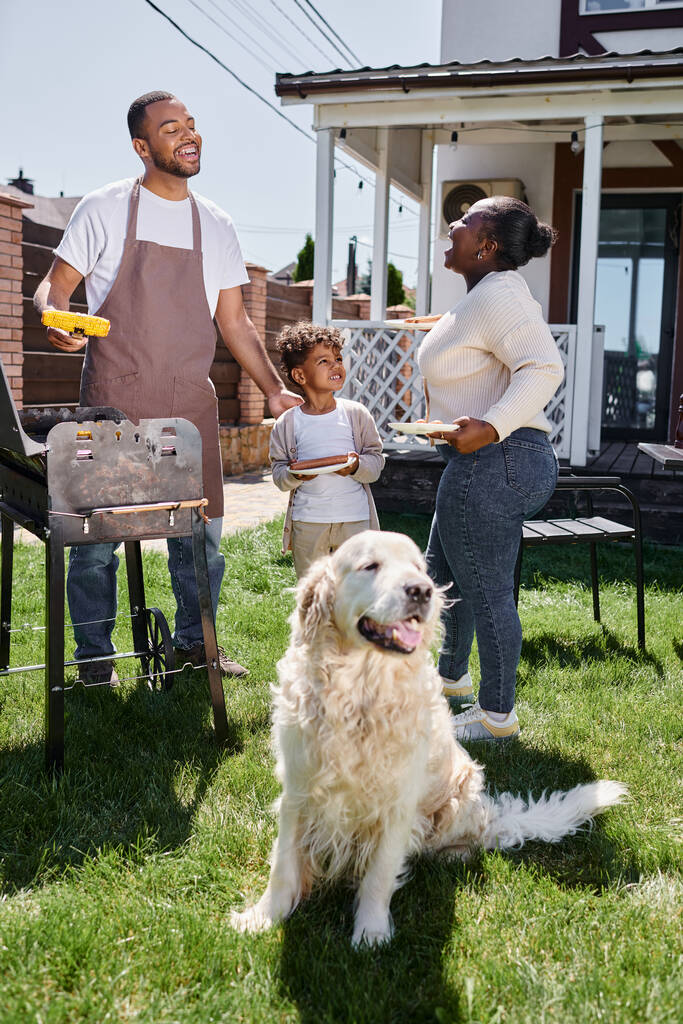 wesoły Afroamerykanin człowiek gotowanie grillowana kukurydza na grill grill w pobliżu psa, żona i syn na podwórku - Zdjęcie, obraz