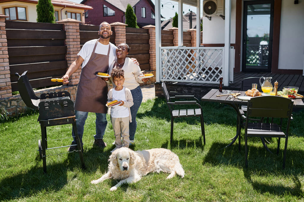 pozytywnych african amerykańskiej rodziny gospodarstwa talerze z grillowanej kukurydzy w pobliżu grilla i pies na podwórku - Zdjęcie, obraz