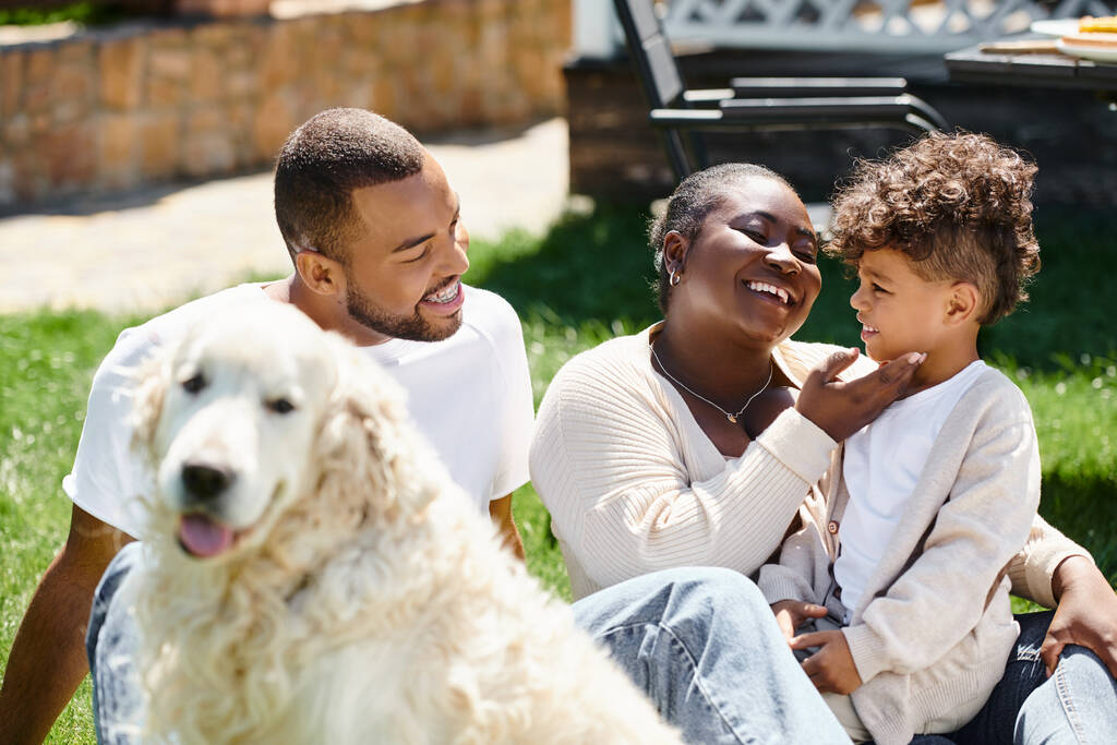 οικογενειακό πορτρέτο της χαρούμενης αφρικανικής οικογένειας χαμογελά και κάθεται στο γκαζόν κοντά στο σκυλί - Φωτογραφία, εικόνα