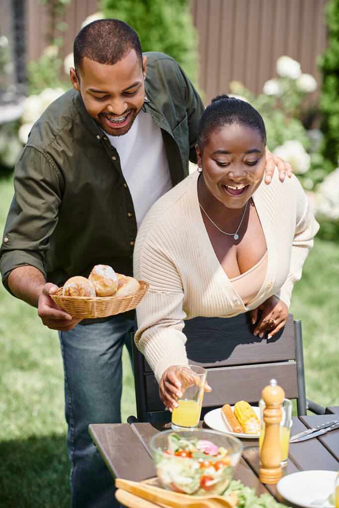 szczęśliwa afrykańska para małżeńska obsługująca stół w ogrodzie, bułki i szklankę soku pomarańczowego - Zdjęcie, obraz