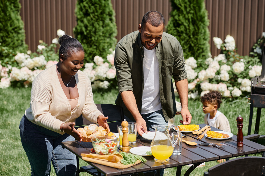 alegres pais americanos africanos servindo mesa no jardim perto do filho brincando no quintal, refeição compartilhada - Foto, Imagem