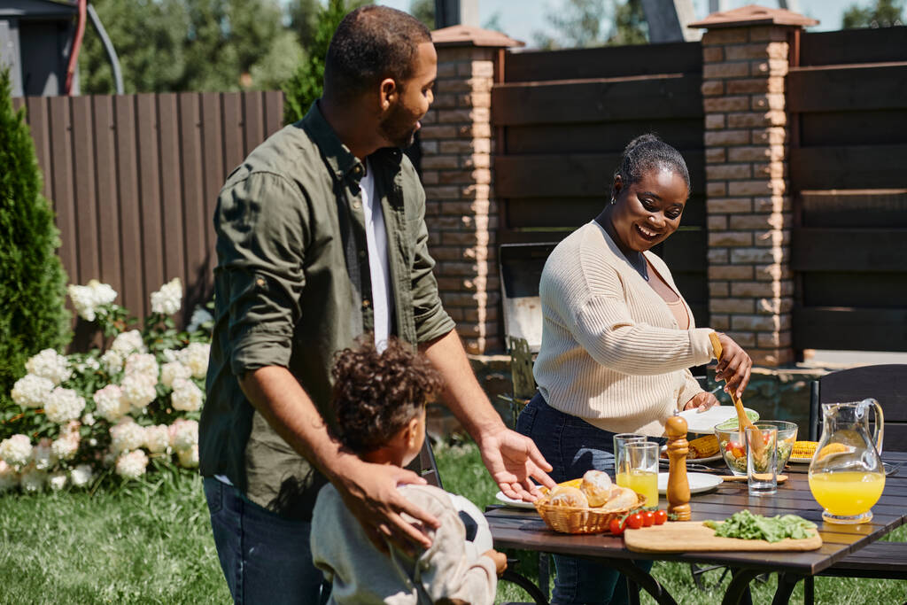幸せなアフリカ系アメリカ人の両親は,裏庭にサッカーボールを持っている息子の近くの庭でテーブルを提供しています - 写真・画像