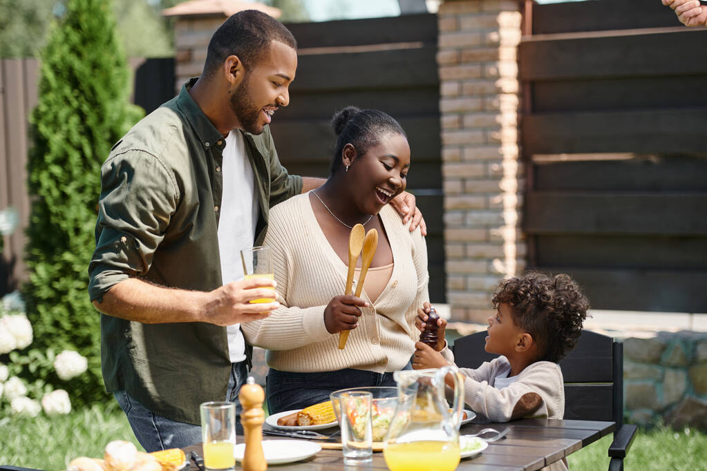 retrato de padres afroamericanos felices y su hijo preparando la mesa para el almuerzo en el jardín en el patio trasero - Foto, imagen