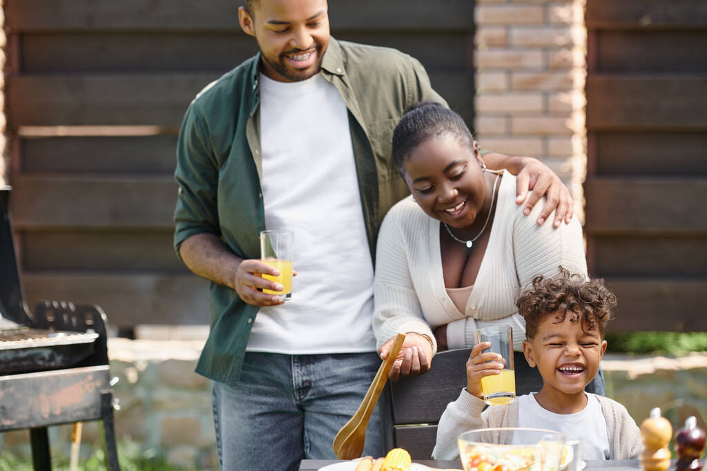 bbqの間に裏庭にオレンジジュースを握っている楽しい息子を見ているアフリカ系アメリカ人の両親 - 写真・画像