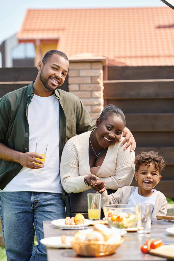 幸せなアフリカ系アメリカ人の両親と息子の肖像画が裏庭で昼食のための庭のテーブルを準備 - 写真・画像