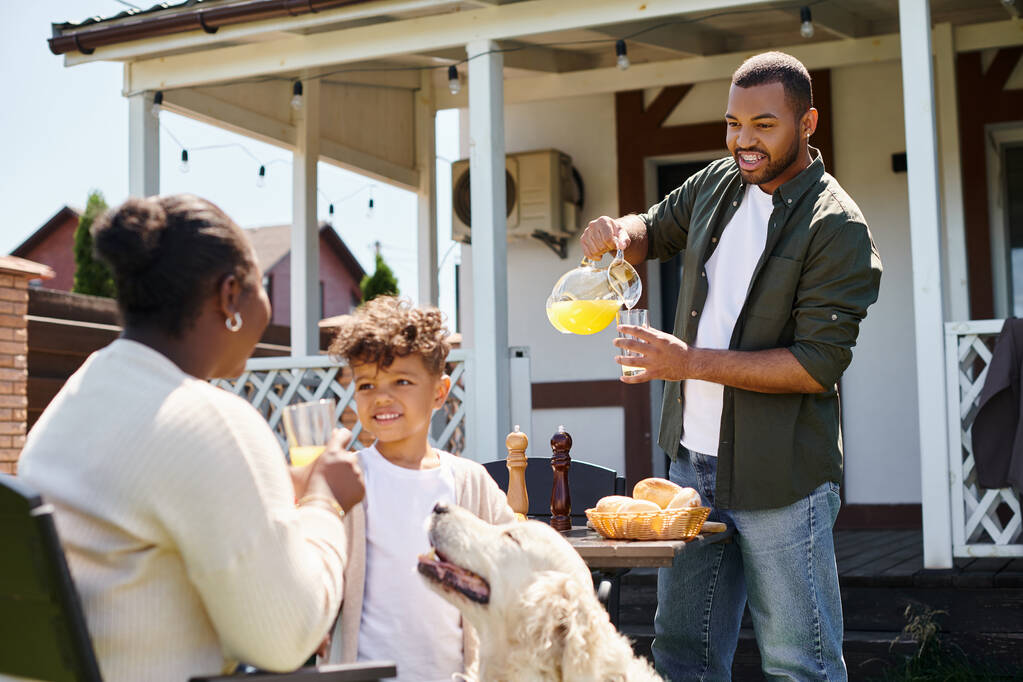 wesoły afrykański amerykański chłopiec uśmiecha się i stoi w pobliżu psa i rodziców podczas rodzinnego grilla na podwórku - Zdjęcie, obraz
