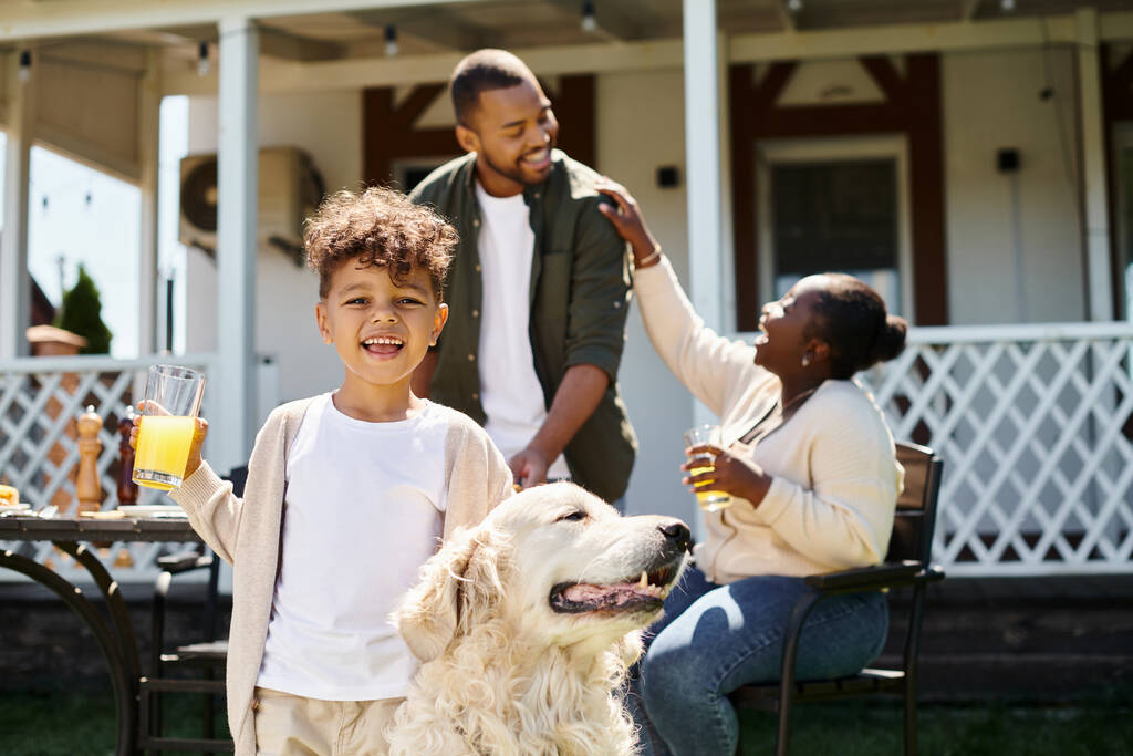 boldog afro-amerikai fiú mosolygós és simogató kutya, miközben egy pohár narancslevet tart a szülők közelében - Fotó, kép