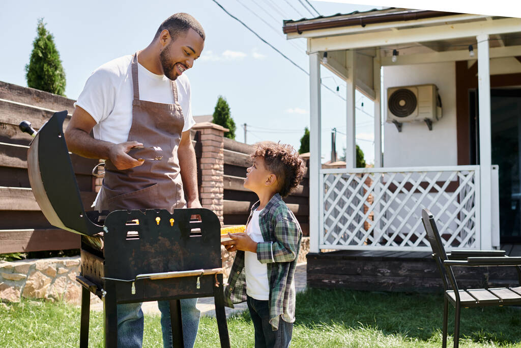 heureux homme afro-américain dans tablier cuisson du maïs sur barbecue grill et en regardant son fils sur la cour - Photo, image