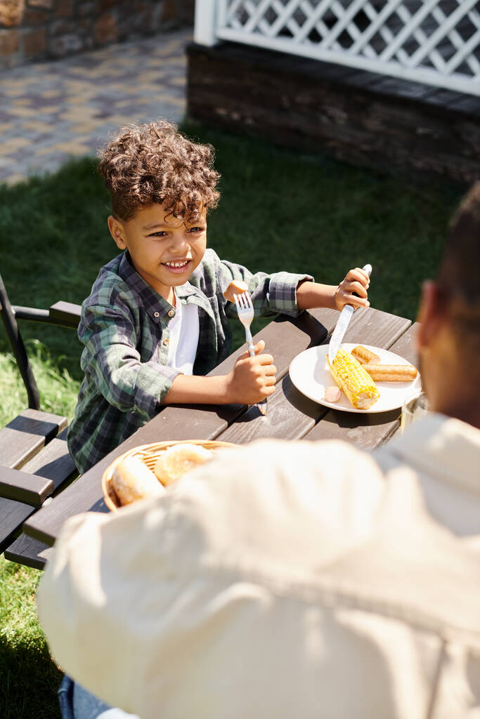 Mutlu kıvırcık Afrikalı Amerikalı çocuk açık havada babaya bakarken sosis yiyip ızgara mısır yiyor. - Fotoğraf, Görsel